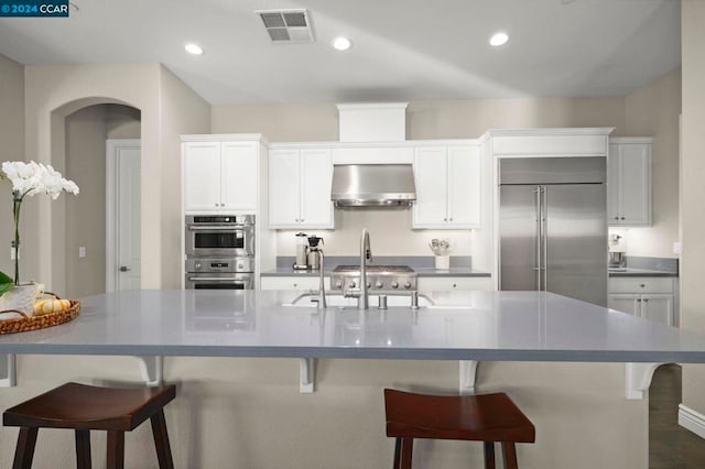 kitchen with wall chimney range hood, white cabinetry, stainless steel appliances, and a large island with sink