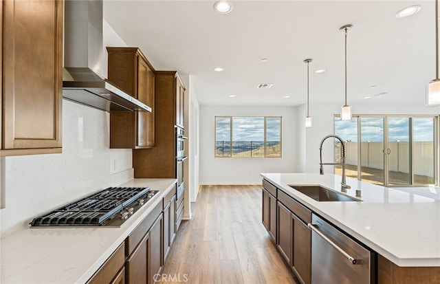 kitchen with a sink, wall chimney range hood, light wood-style floors, stainless steel appliances, and a kitchen island with sink