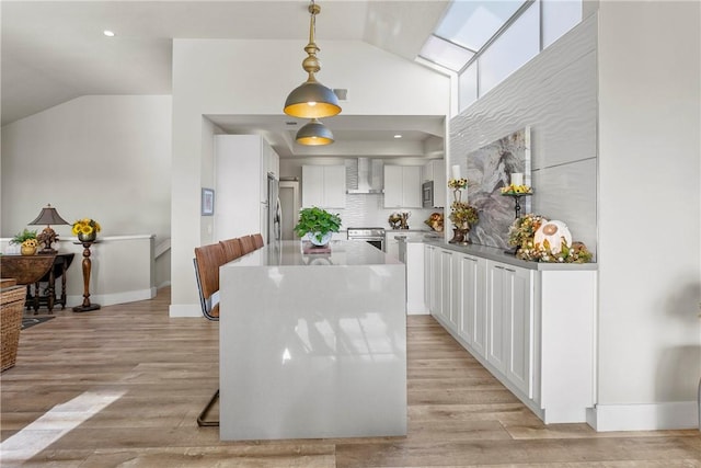 kitchen with wall chimney exhaust hood, stainless steel appliances, hanging light fixtures, white cabinets, and light wood-type flooring