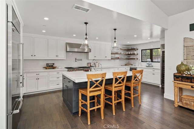 kitchen with a center island with sink, wall chimney exhaust hood, stainless steel built in refrigerator, and dark wood-type flooring