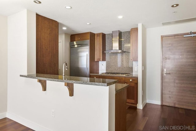 kitchen with a breakfast bar, dark wood-type flooring, wall chimney range hood, kitchen peninsula, and stainless steel appliances