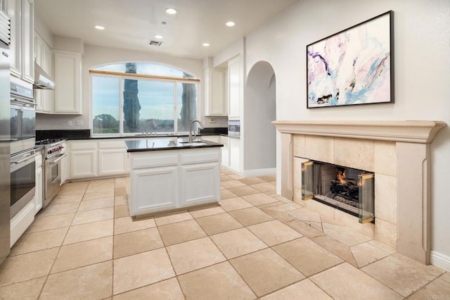 kitchen featuring stainless steel range, a kitchen island with sink, white cabinets, a tiled fireplace, and sink