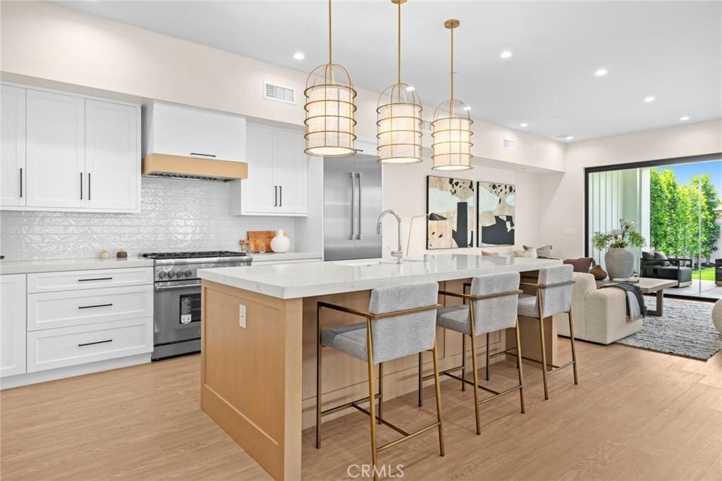 kitchen with pendant lighting, an island with sink, white cabinets, high end range, and light hardwood / wood-style floors