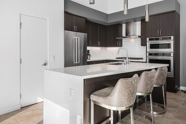 kitchen featuring a breakfast bar, appliances with stainless steel finishes, hanging light fixtures, an island with sink, and wall chimney exhaust hood