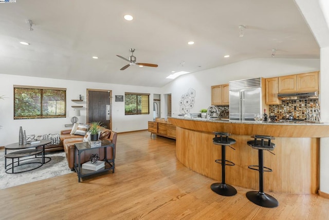 kitchen featuring light brown cabinetry, vaulted ceiling, light hardwood / wood-style flooring, a kitchen breakfast bar, and built in fridge