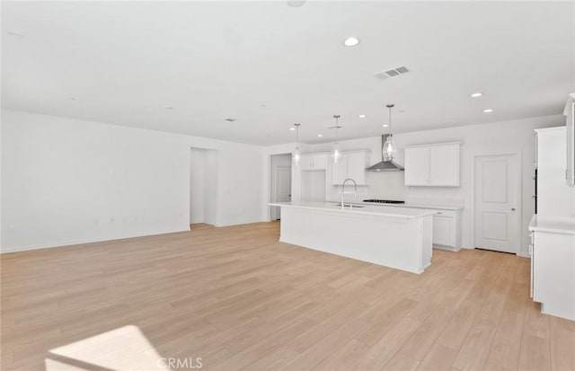 kitchen featuring an island with sink, white cabinets, wall chimney range hood, decorative light fixtures, and light hardwood / wood-style floors