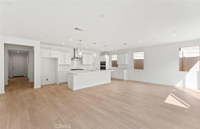 kitchen with sink, white cabinetry, decorative light fixtures, wall chimney range hood, and a kitchen island with sink