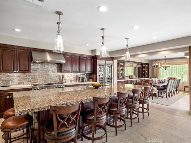 kitchen featuring decorative backsplash, a large island with sink, wall chimney exhaust hood, a breakfast bar area, and pendant lighting