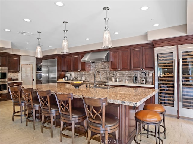 kitchen featuring beverage cooler, a large island with sink, wall chimney range hood, stainless steel appliances, and a breakfast bar