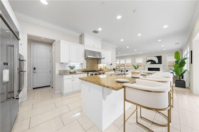 kitchen featuring white cabinetry, light stone counters, ventilation hood, a breakfast bar area, and a center island with sink