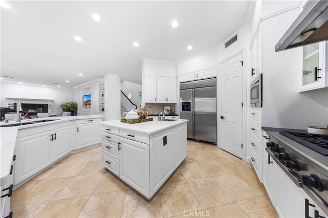 kitchen with built in appliances, white cabinets, an island with sink, and range hood