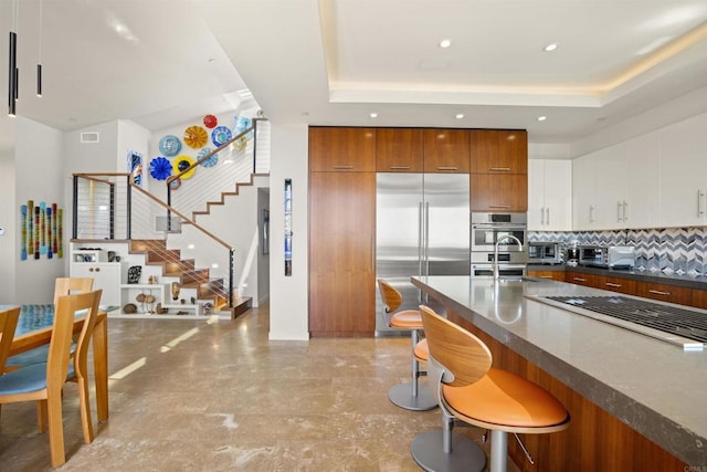 kitchen featuring a raised ceiling, decorative backsplash, appliances with stainless steel finishes, white cabinetry, and a breakfast bar area