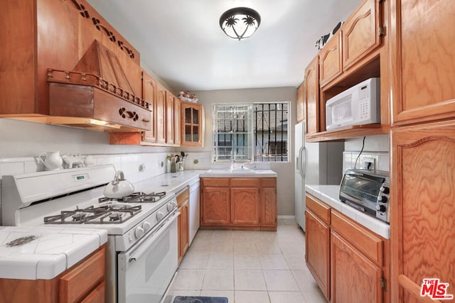 kitchen with custom range hood, decorative backsplash, white appliances, light tile patterned floors, and sink