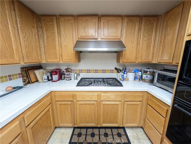 kitchen with tile countertops, decorative backsplash, light tile patterned floors, black oven, and stainless steel gas cooktop