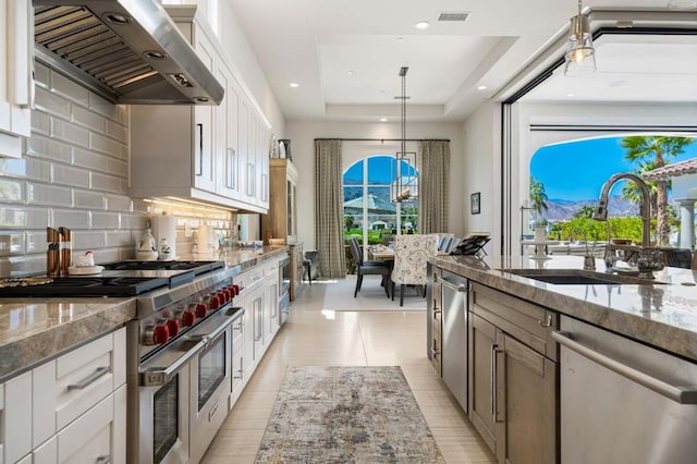 kitchen featuring wall chimney range hood, light stone countertops, appliances with stainless steel finishes, decorative light fixtures, and white cabinetry