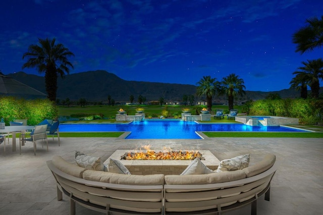 pool at night with an in ground hot tub, an outdoor living space with a fire pit, pool water feature, a mountain view, and a patio