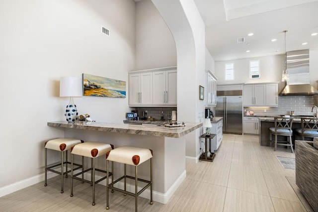 kitchen with a kitchen bar, kitchen peninsula, wall chimney range hood, pendant lighting, and stainless steel built in fridge