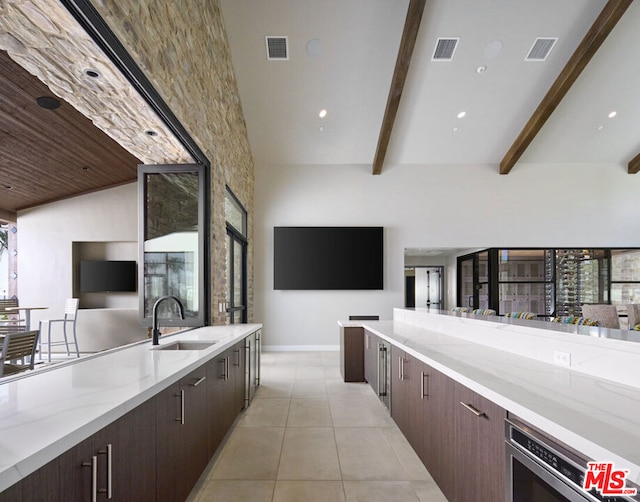 kitchen featuring dark brown cabinetry, beamed ceiling, sink, light stone counters, and light tile patterned floors