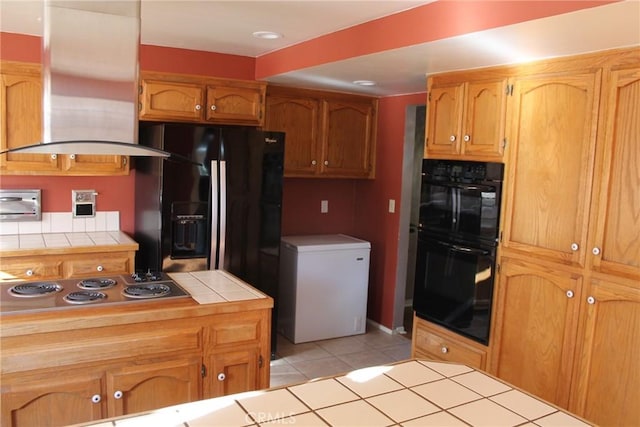 kitchen featuring brown cabinetry, island exhaust hood, light tile patterned floors, black appliances, and tile counters