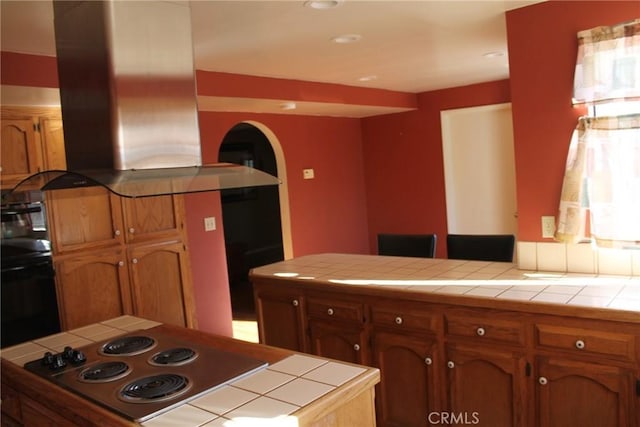 kitchen with brown cabinets, tile countertops, island range hood, and stainless steel electric cooktop