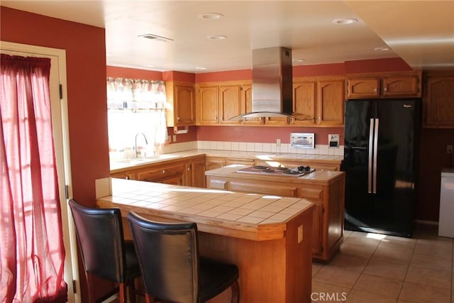 kitchen with tile countertops, island range hood, black fridge with ice dispenser, a sink, and stainless steel cooktop