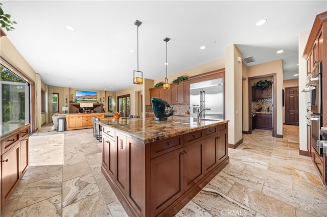 kitchen featuring a large island with sink, dark stone countertops, built in refrigerator, backsplash, and pendant lighting