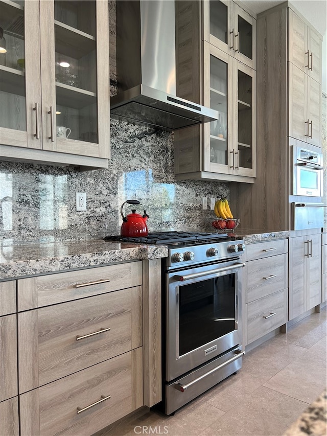 kitchen with wall chimney range hood, light brown cabinets, decorative backsplash, light stone countertops, and appliances with stainless steel finishes