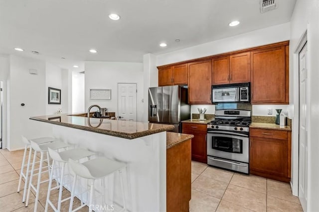 kitchen with a center island with sink, dark stone countertops, light tile patterned floors, a kitchen bar, and stainless steel appliances