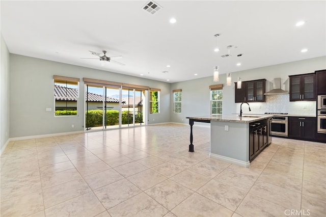 kitchen with appliances with stainless steel finishes, a kitchen breakfast bar, dark brown cabinets, a center island with sink, and wall chimney range hood