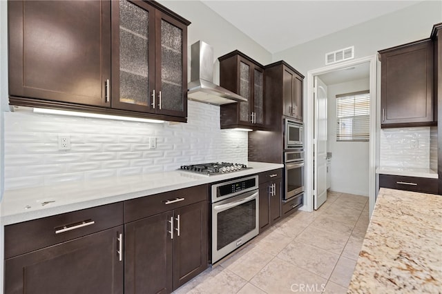 kitchen with wall chimney exhaust hood, stainless steel appliances, backsplash, and light stone countertops