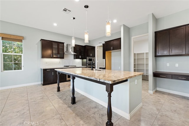 kitchen with light stone countertops, decorative light fixtures, dark brown cabinetry, a kitchen island with sink, and wall chimney range hood