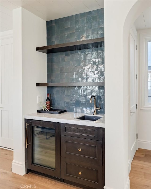 bar with wine cooler, sink, dark brown cabinets, light wood-type flooring, and decorative backsplash