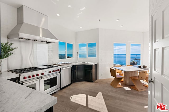 kitchen featuring appliances with stainless steel finishes, a water view, light stone counters, light wood-type flooring, and wall chimney range hood