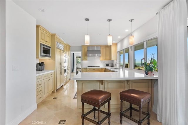 kitchen featuring light countertops, stainless steel microwave, a sink, wall chimney range hood, and a peninsula