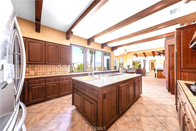 kitchen with tile counters, an island with sink, tasteful backsplash, sink, and stainless steel refrigerator