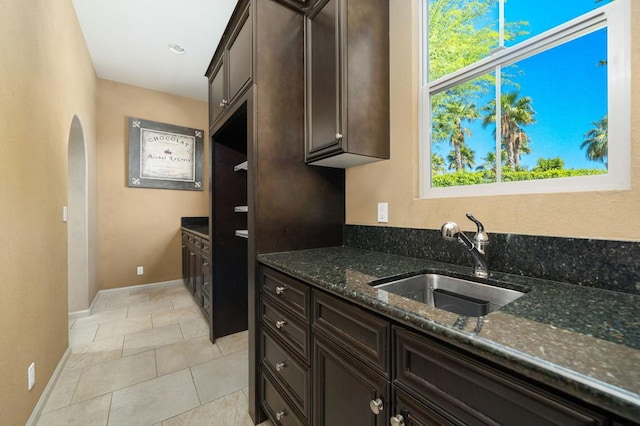 kitchen with dark brown cabinets, dark stone countertops, light tile patterned floors, and sink