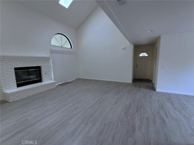 unfurnished living room featuring a skylight, a brick fireplace, high vaulted ceiling, and light hardwood / wood-style flooring