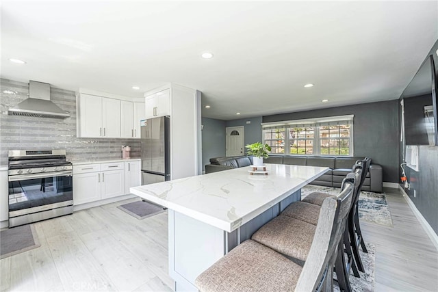 kitchen with white cabinetry, light stone counters, stainless steel appliances, light wood-type flooring, and wall chimney range hood