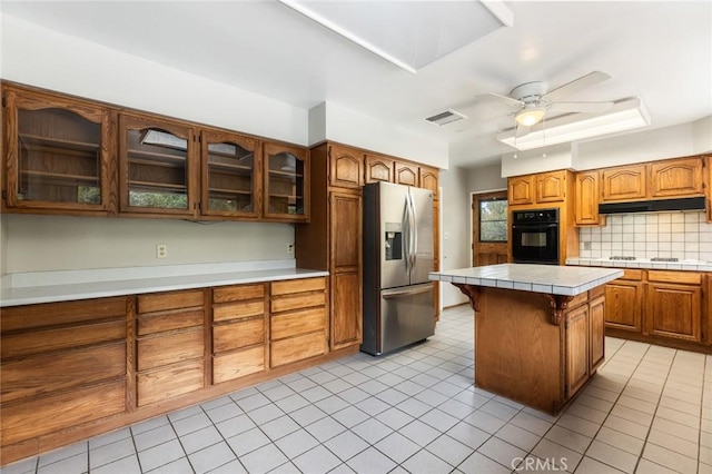 kitchen featuring stainless steel refrigerator with ice dispenser, ceiling fan, black oven, tile counters, and a kitchen island