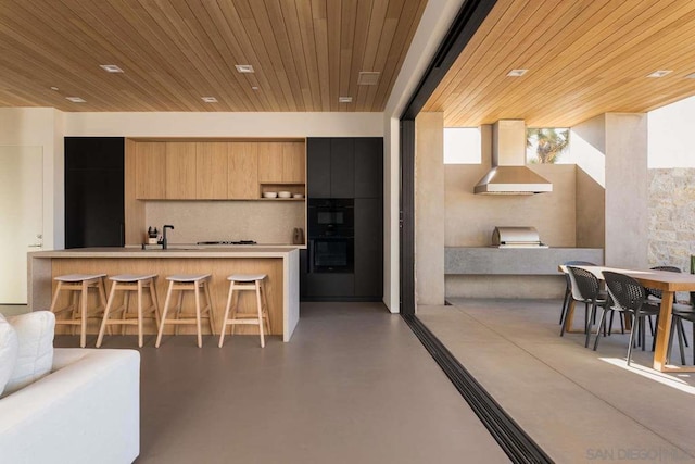 kitchen with a kitchen breakfast bar, wood ceiling, tasteful backsplash, wall chimney exhaust hood, and black double oven