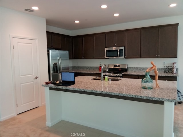 kitchen with appliances with stainless steel finishes, light stone counters, dark brown cabinets, and an island with sink