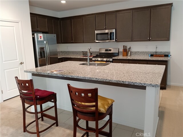 kitchen with light tile patterned floors, a kitchen island with sink, light stone countertops, dark brown cabinetry, and stainless steel appliances