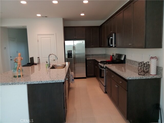 kitchen with an island with sink, stainless steel appliances, sink, light stone countertops, and dark brown cabinetry