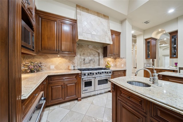 kitchen with light stone counters, backsplash, range with two ovens, custom exhaust hood, and sink