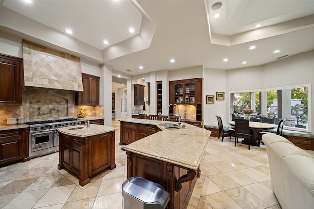 kitchen featuring a tray ceiling, high end stove, sink, decorative backsplash, and a center island with sink