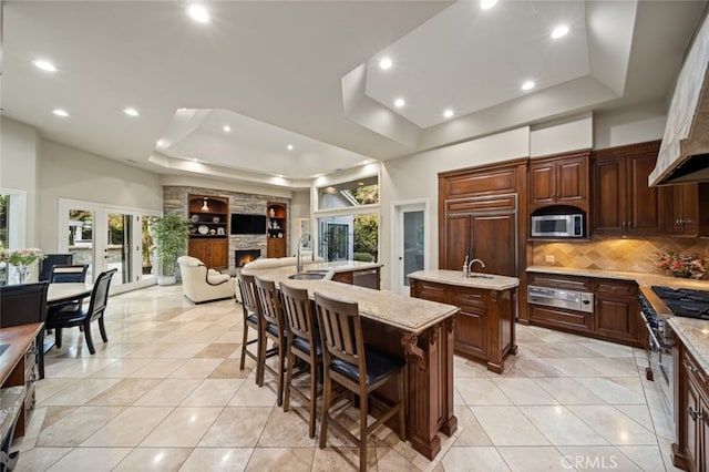 kitchen featuring light tile patterned floors, a stone fireplace, a kitchen island with sink, and a tray ceiling