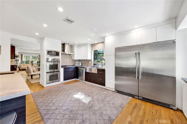 kitchen with appliances with stainless steel finishes, decorative backsplash, white cabinets, light wood-type flooring, and wall chimney range hood