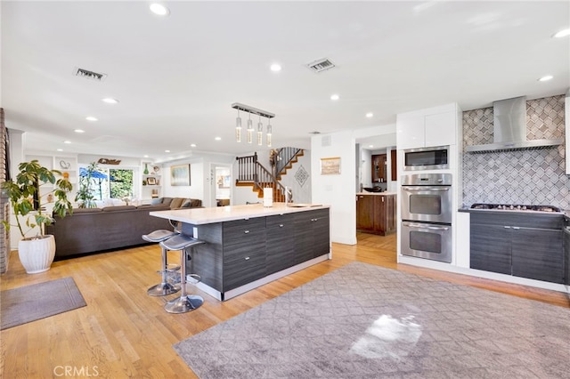 kitchen with light hardwood / wood-style floors, wall chimney exhaust hood, an island with sink, and tasteful backsplash