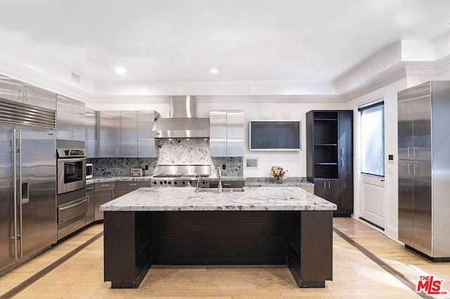 kitchen with a kitchen island with sink, light hardwood / wood-style flooring, wall chimney range hood, and appliances with stainless steel finishes