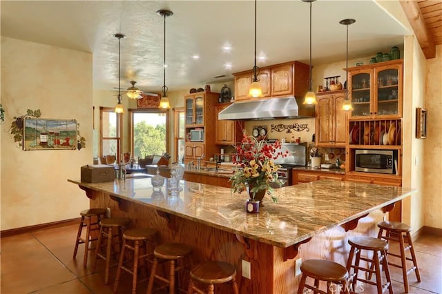 kitchen with under cabinet range hood, light stone countertops, appliances with stainless steel finishes, glass insert cabinets, and brown cabinets